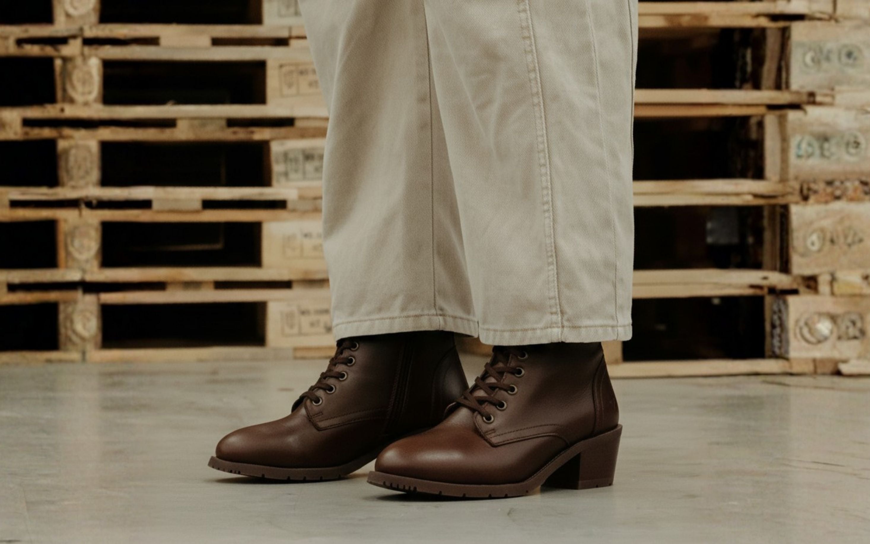 Woman wearing Xena Vega steel-toe safety boots in chestnut brown leather with wide-leg pants, standing among warehouse pallets